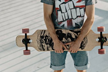 Erkek eller ahşap bir kaykayı tutar. Ellerinde kaykay tutan, doğanın arka planında kayan genç bir adamın yakın çekimi. Bedava gevşetici kaykay konsepti. hipster tarzı