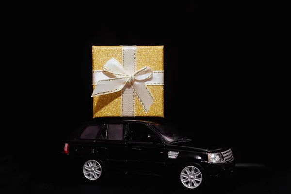 black toy car carries a gift. Gold box with ribbon. Concept of Christmas mood and preparation for the celebration. Discounts. Present. Black Friday. 2022. Seasonal sale, big discounts