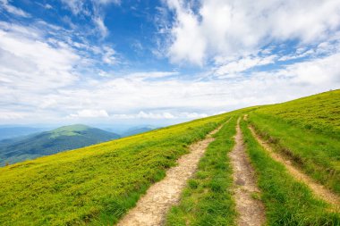 Güneşli bir günde dağ manzarası. Çimenli çayırlarda iz var. Güzel bir seyahat manzarası. Uzaklarda, mavi gökyüzünün altında çarpıcı bulutlu bir tepe..