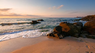 Güneşin doğuşunda kumlu sahilde kocaman taşlar. Sabah vakti Karadeniz 'de harika bir kadife sezonu tatili. Durgun dalgalar, İspanya kıyılarını yıkıyor. Ufkun üstündeki mavi gökyüzünde parlayan bulutlar