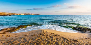 Kumsalda sakin bir sabah manzarası. Harika bir yaz tatili geçmişi. Sakin dalgalar kıyıları yıkar. Mavi gökyüzü ve sabah ışığında parlayan bulutlar. Kuma basıyor. Taşın üzerindeki yosun