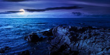 Geceleri kayalık deniz kıyısında. Yaz tatili konsepti. Güzel doğa manzarası, mavi gökyüzünde bulutlar, ufukta dolunay ışığı.