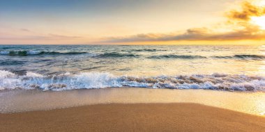 Güneşin doğuşunda kumlu sahil. Güzel bir deniz manzarası. Sakin dalgalar kıyıları yıkıyor. Sabah ışığında parlayan bulutlar. Yaz tatili ve dinlenme konsepti