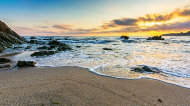 Gün doğumunda boş bir sahil. Denizin güzel manzarası. Dalgalar sahildeki kum ve taşları yıkıyor. Sabah ışığında parlayan bulutlar. sakin bir yaz tatili ve seyahat yeri konsepti