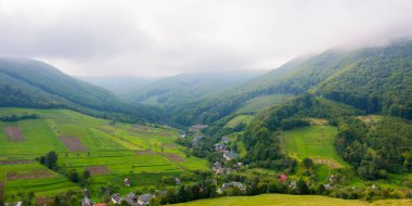 Karpatya kırsal arazisi. Yeşil alanlar ve tepelerde ekilebilir. Vadideki küçük bir köy. Parlak bulutlu gökyüzünün altındaki ormanlı dağ