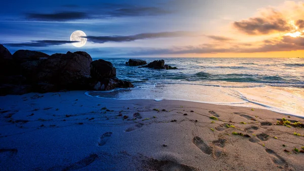 day and night time change concept above huge stones on the sandy beach at twilight. wonderful velvet season vacation. clouds on the blue sky with sun and moon above horizon. waves washing the shore
