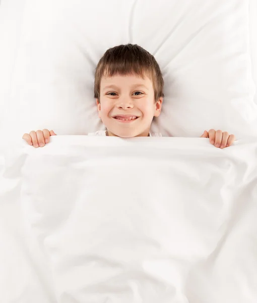 7 years old boy hiding in bed under a white blanket or coverlet Stock