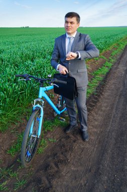 Bisikletli bir iş adamı yeşil bir çimenlikte kol saatine bakıyor. Özgürlük, tatil ya da serbest çalışma konsepti. Güzel bahar doğası.