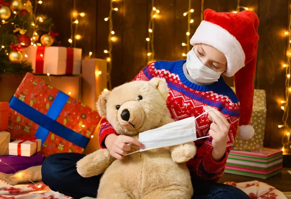 サンタのヘルパーとしての子供の女の子は 新年の装飾にポーズ ウイルスに対する保護面マスクを着て コロナウイルス 彼女はおもちゃの熊にマスクをつけた 休日のライトや贈り物の多くは おもちゃとエレガントなクリスマスツリー — ストック写真