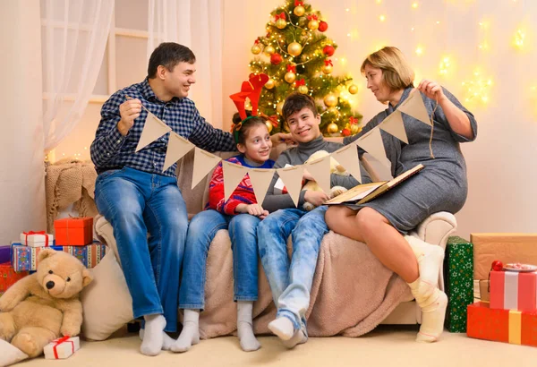 寒假的概念 家庭在室内摆设与新年或圣诞节装饰 坐在沙发上 展示节日彩旗的孩子和家长 假日灯具和礼物 — 图库照片