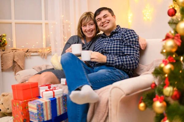 男人和女人坐在沙发上 摆出新年或圣诞节的姿势 他们用白色杯子喝茶或喝咖啡假日的灯和礼物 装饰着玩具的圣诞树 — 图库照片