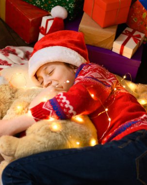 Yeni yılda ya da Noel süslemesinde uyuyan kız çocuğu. Tatil ışıkları ve hediyeler, oyuncaklarla süslenmiş Noel ağacı. Kırmızı bir kazak ve Noel Baba şapkası giyiyor.