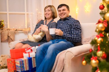 Kanepede oturan erkek ve kadın yeni yılda ya da Noel süslemesinde poz veriyorlar. Konuşuyorlar ve çay içiyorlar ya da beyaz bardaklardan kahve içiyorlar tatil ışıkları ve hediyeler, oyuncaklarla süslenmiş Noel ağacı..