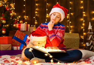 Yeni yılda kitap okuyan bir çocuk ya da noel süsü. Tatil ışıkları ve hediyeler, oyuncaklarla süslenmiş Noel ağacı. Kırmızı bir kazak ve Noel Baba şapkası giyiyor.