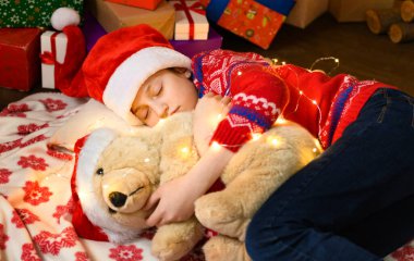 Yeni yılda ya da Noel süslemesinde uyuyan kız çocuğu. Tatil ışıkları ve hediyeler, oyuncaklarla süslenmiş Noel ağacı. Kırmızı bir kazak ve Noel Baba şapkası giyiyor.