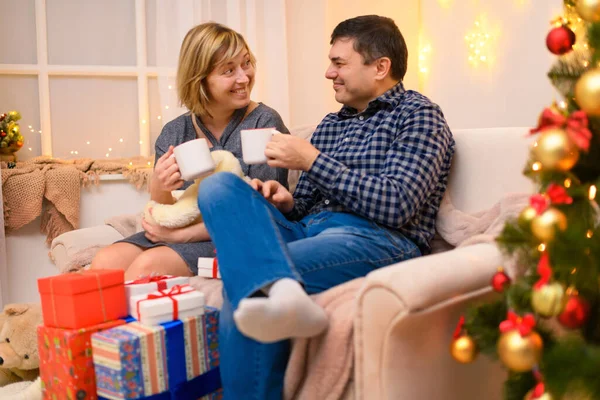 男人和女人坐在沙发上 摆出新年或圣诞节的姿势 他们笑着喝着茶或白杯中的咖啡假日的灯和礼物 装饰着玩具的圣诞树 — 图库照片