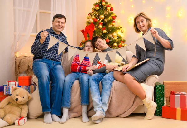 寒假的概念 家庭在室内摆设与新年或圣诞节装饰 坐在沙发上 展示节日彩旗的孩子和家长 假日灯具和礼物 — 图库照片