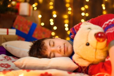 Yeni yılda ya da Noel süslemesinde uyuyan ergen çocuk. Tatil ışıkları, hediyeler ve oyuncaklarla süslenmiş Noel ağacı..