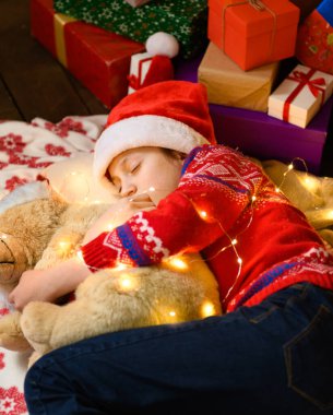 Yeni yılda ya da Noel süslemesinde uyuyan kız çocuğu. Tatil ışıkları ve hediyeler, oyuncaklarla süslenmiş Noel ağacı. Kırmızı bir kazak ve Noel Baba şapkası giyiyor.