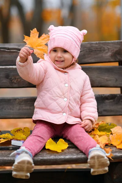 秋の都市公園のベンチに座ってカエデの葉で遊んでいる子供の少女の肖像画 美しい自然 黄色の葉を持つ木 — ストック写真