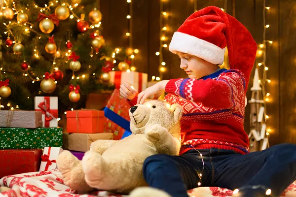 孩子们在新年或圣诞节装扮中摆姿势 节日灯火通明 礼物繁多 圣诞树上放着精美的玩具 这个女孩穿着一件红色的毛衣 戴着一顶圣诞老人的帽子 她在和熊玩耍 — 图库照片