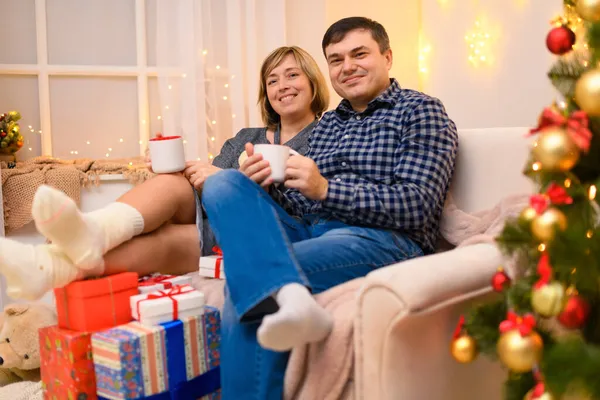 男人和女人坐在沙发上 摆出新年或圣诞节的姿势 他们用白色的杯子喝茶或喝咖啡假日的灯和礼物 装饰着玩具的圣诞树 — 图库照片