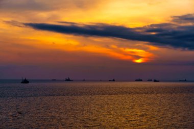 Denizde ve gemilerde güzel bir gün batımı. Güneş bulutların arasından parlıyor ve dalgalara yansıyor.