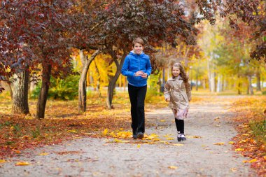 Çocuklar sonbahar parkında yol boyunca koşar. Oynuyor ve gülüyorlar, çok eğleniyorlar. Çevreleri güzel doğa ve sarı ve kırmızı yapraklı ağaçlarla çevrili.