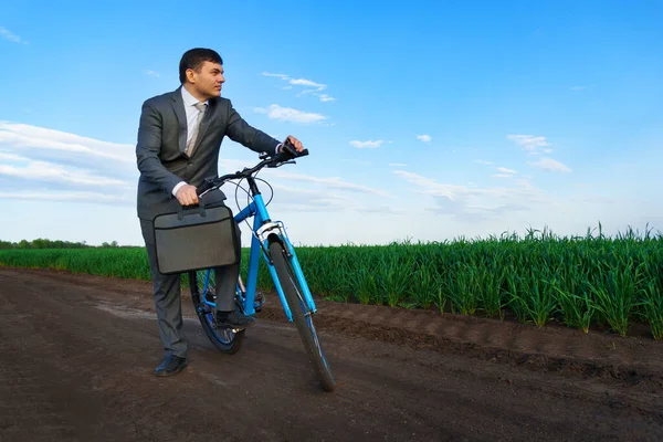 ビジネスコンセプト ビジネスマンは緑の芝生のフィールドに自転車に乗って ビジネススーツを着て 彼はブリーフケースと書類を持っています — ストック写真