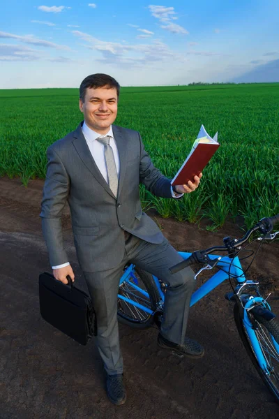 사업가가 풀밭에서 자전거를 다니면서 가방에서 일기장을 꺼내서 양복을 — 스톡 사진