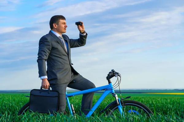 ビジネスコンセプト ビジネスマンは緑の芝生のフィールドに自転車に乗って 双眼鏡を通して距離を調べ ビジネススーツを着て 彼はブリーフケースと書類を持っています — ストック写真