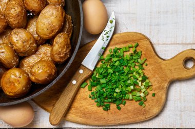 Kızartma tavasında kızarmış patates, sarımsak ve yeşil soğan, çiftlik yumurtası ve tuz, beyaz ahşap arka planda kırsal tarzda pişmiş ve çiğ yemek.