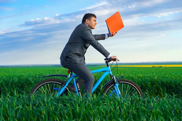 一位拥有红色文件夹以获取报告或文件的商人骑着自行车穿过绿草场 穿着西装 夏天的美丽自然 商业理念 — 图库照片
