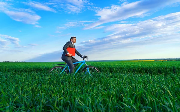 レポートや書類のための赤いフォルダを持つビジネスマンは 緑の芝生のフィールドを介して自転車に乗る ビジネススーツに身を包んだ 春の美しい自然 ビジネスコンセプト — ストック写真