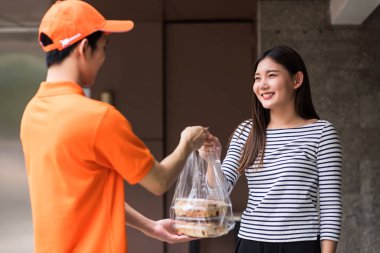 Turuncu üniformalı teslimatçı apartman dairesinin veya apartmanın önündeki gülümseyen kadına tatlı ekmek fırını veriyor. Covid-19 delta salgını sırasında internetten yemek sipariş etmek için yeni bir normal.