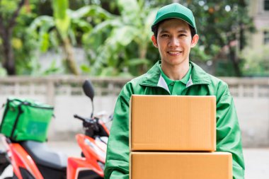 Gülümseyen kurye yeşil ceketli kurye bulanık motosiklet arkaplanlı paket kutuları veriyor. Cep telefonu uygulaması ile online alışveriş. Ev teslimatı covid-19 'u önlemek için.