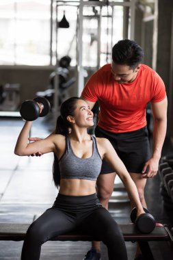Spor salonundaki Asyalı güzel bir kadına pazı egzersizi için Dumbbell 'ı çalıştıran antrenör ya da erkek arkadaş. Mutlu Asyalı çift vücut geliştirme ve sağlıklı yaşam için çalışıyor..