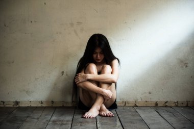 Genç Asyalı kadın üzgün ve stresli bir şekilde ahşap zeminde oturuyor. Seksi fahişe. Boş harabe odasında üzgün. Pencere kenarında açık renk bir gölge var..