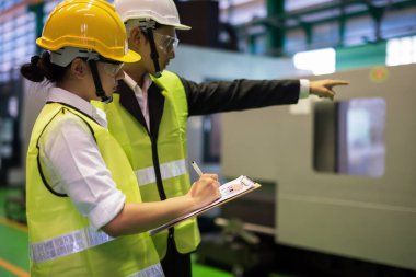 Bayan fabrika müfettişi, denetleme müdürü parmağını gösterirken mikro çip makinesini inceledikten sonra taslak taslağı not alıyor. CNC otomatik torna makineleriyle sanayi üretimi. Kalite güvencesi