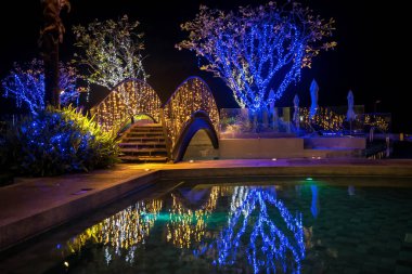Lüks yüzme havuzundaki köprü ve ağaçlar üzerindeki süslemeleri aydınlatın. Geceleri modern tatil köylerinde ya da otellerde su yansıtılır..