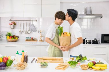 Yakışıklı Asyalı eşcinsel, mutfakta yemek dağıtırken ortağını öpüyor. Evde mutlu bir LGBT yaşantısı. Sevgililer Günü 'nde salata hazırlamak için sağlıklı beslenen aynı cinsiyetten eşcinsel aile..