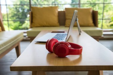 Kanepe masasındaki dizüstü bilgisayarın yanında kırmızı kablosuz kulaklık ya da cep telefonu. Müzik ve dijital ekipman kavramı