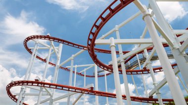 red roller coaster in a amusement park with some loopings against blue sky and white cloud.