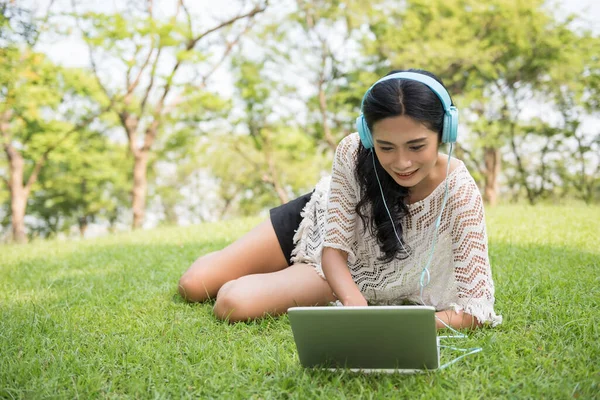 Joyful asian smiling girl sitting on lawn with laptop, studying and listening music in spring