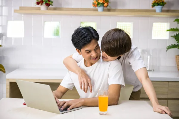 Young LGBT Asian gay man hug his partner who work on laptop computer from behind. Happy couple together at home. Same sex family.
