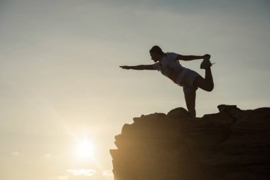 Silhouette athlete sport woman perform yoga exercise on top of hill during sunset after climbing to mountain summit peak. She stretching arms, hands, and legs. Exstreme sport and healthy lifestyle.