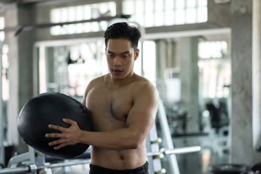 Fit Asian muscle man with nake torso swing abdomen ball at fitness gym. Man exercise by swinging hard medicine ball in sport club. Bodybuilding and healthy lifestyle concept.