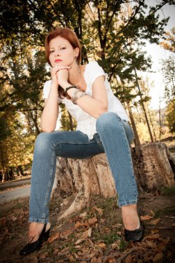 Beutiful redhead girl in a white t-shirt and jeans sitting on a tree stump clipart