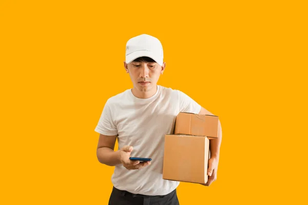 Young Asian delivery man wearing cap with white blank t-shirt holding smartphone and parcel post box isolated on yellow background. express delivery service concept.