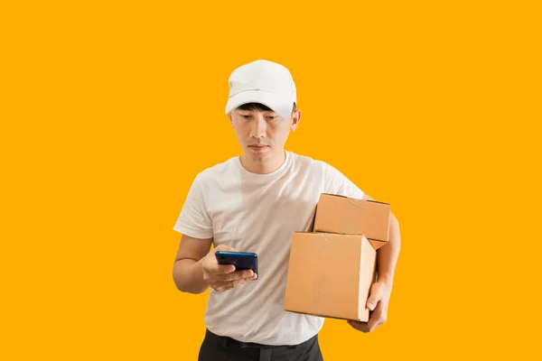 Young Asian delivery man wearing cap with white blank t-shirt holding smartphone and parcel post box isolated on yellow background. express delivery service concept.